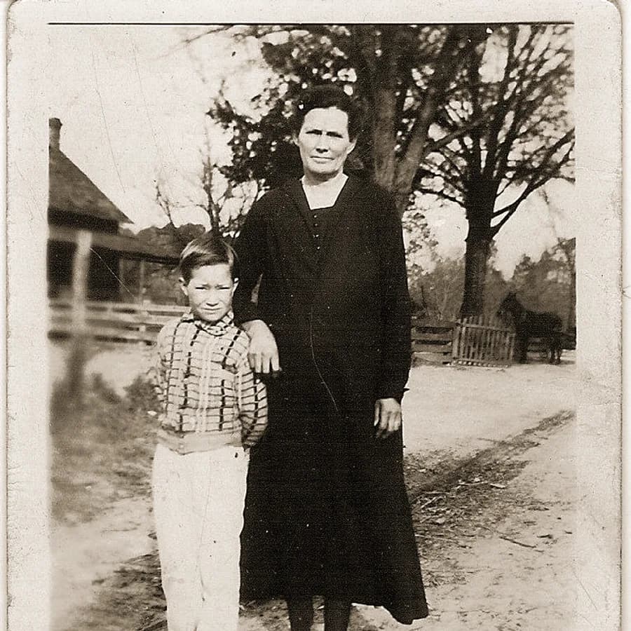 Mulher de vestido escuro e um menino numa estrada de terra, diante de uma casa